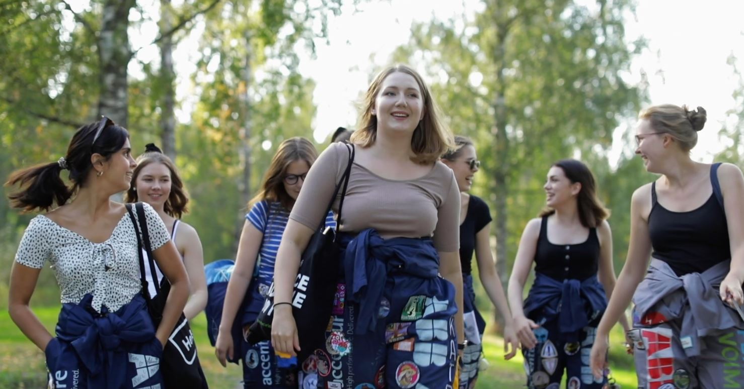 Joukko opiskelijoita kävelee opiskelijahaalarit yllään kesäisessä maisemassa, taustalla koivuja.